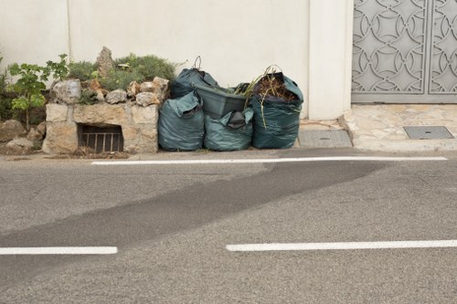 The image depicts three large, dark green plastic rubbish bags filled with waste, positioned on the edge of a paved road in a residential area. The bags are placed near a white wall with a small garden bed to the left, containing some green plants and rocks. Behind the bags, there is a ground-level vent or drainage grille. To the right, part of a decorative grey metal gate or panel is visible. The road surface is grey with white lane markings, and the scene appears to be outdoors during daytime with natural lighting. The setting suggests a local rubbish removal context, possibly in Seven Sisters, aligning with a professional waste management service such as Office Clearance Seven Sisters.