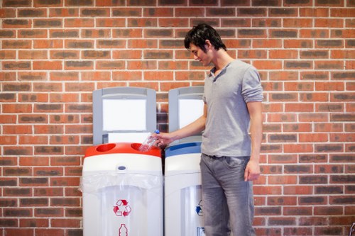 A young man with dark hair, wearing a light grey long-sleeved shirt and grey trousers, stands outdoors on a paved area adjacent to a red brick wall, placing a plastic bottle into a recycling bin marked with a red recycling symbol. Next to him are two large, cylindrical rubbish containers with flat tops; one container is white with a red recycling symbol, and the other is white with a blue symbol, both placed side by side against the brick wall. The scene suggests a waste management activity related to rubbish removal or recycling collection, typical in urban or suburban settings near residential or commercial premises in London or nearby areas. Office Clearance Seven Sisters offers services related to rubbish clearance, and this image exemplifies proper disposal or recycling practices in the local environment.