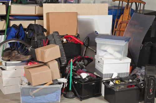 A cluttered indoor space with a large assortment of discarded household items and packaging, including several cardboard boxes, some empty and others filled with various objects, stacked haphazardly. There is a black plastic crate and a transparent plastic container among the items, which contain textiles and smaller miscellaneous objects. A bicycle wheel is partially visible on the left, resting against the pile. In the background, there are shelves with books and storage boxes, and a few pieces of furniture can be seen, indicating this may be a room or a garage awaiting clearance. The scene is well-lit and shows typical signs of rubbish accumulation suitable for professional clearance services, such as those offered by Office Clearance Seven Sisters in the London area, within the N15 postcode. The arrangement suggests ongoing transport or disposal preparation, with various materials and containers unsorted, ready for removal. The environment appears functional and typical of a property pending or undergoing clearance, emphasizing the need for professional rubbish disposal services in the Seven Sisters district.