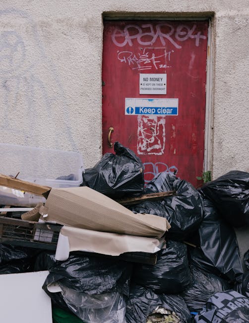 An outdoor urban scene featuring a large, weathered red metal waste container positioned against a dark green wall on the left and a light grey concrete wall on the right. The container appears slight