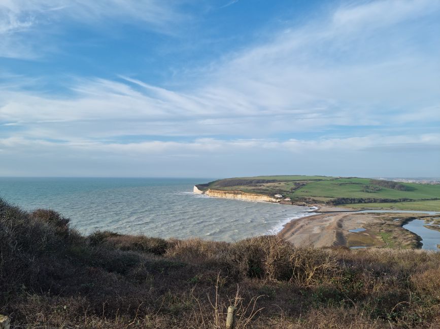 A wide coastal landscape under a clear blue sky, featuring a stretch of grassy terrain in the foreground leading towards a cluster of white chalk cliffs in the background. The cliffs rise sharply from