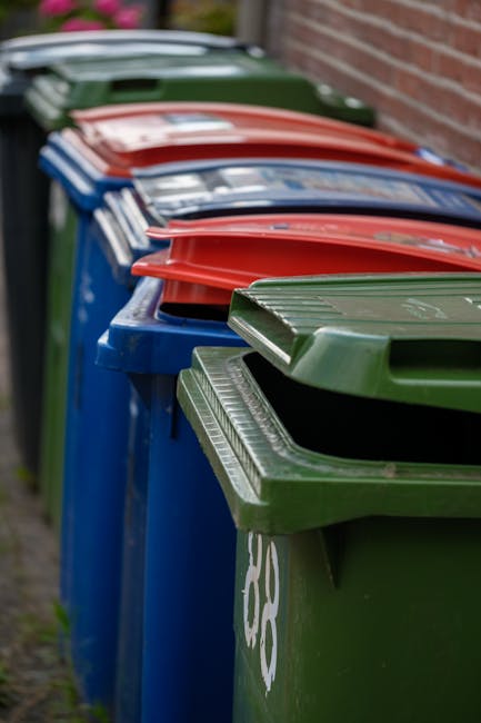A row of wheelie bins arranged along a pavement, with the foreground bin being green and featuring a closed lid with horizontal grooves. Behind it, there are additional bins in blue, red, and dark gre