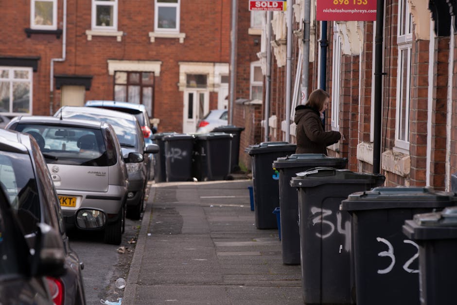 A row of wheelie bins arranged along a pavement, with the foreground bin being green and featuring a closed lid with horizontal grooves. Behind it, there are additional bins in blue, red, and dark gre