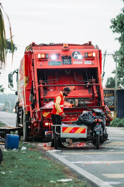 A street scene featuring a red industrial waste collection vehicle parked on the roadside during daylight hours. The vehicle's rear loading compartment is open, revealing mechanical components and a c